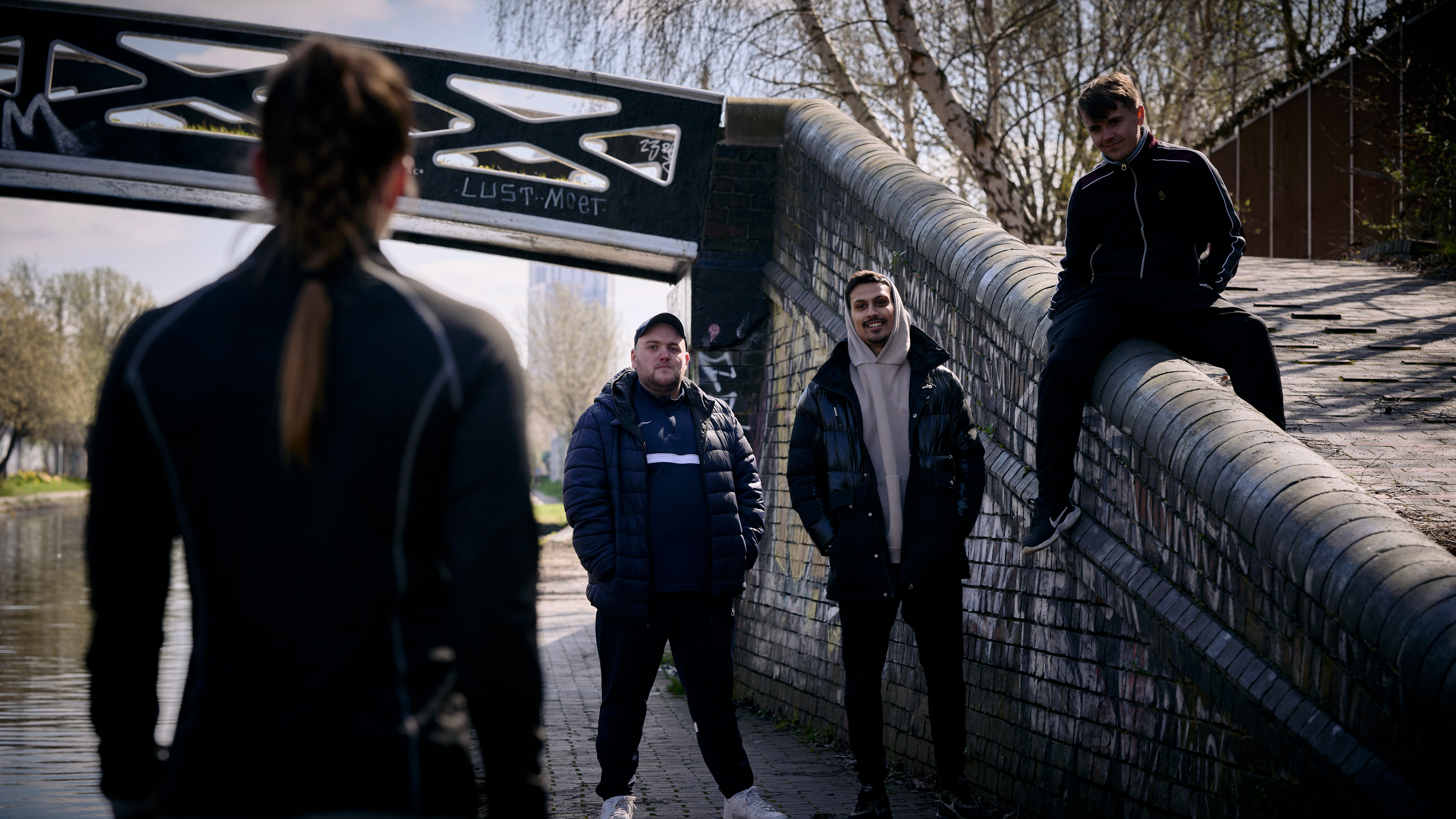 Still from 'I'm Tired Of This' video - a woman on a canal path stops in front of a group of men.
