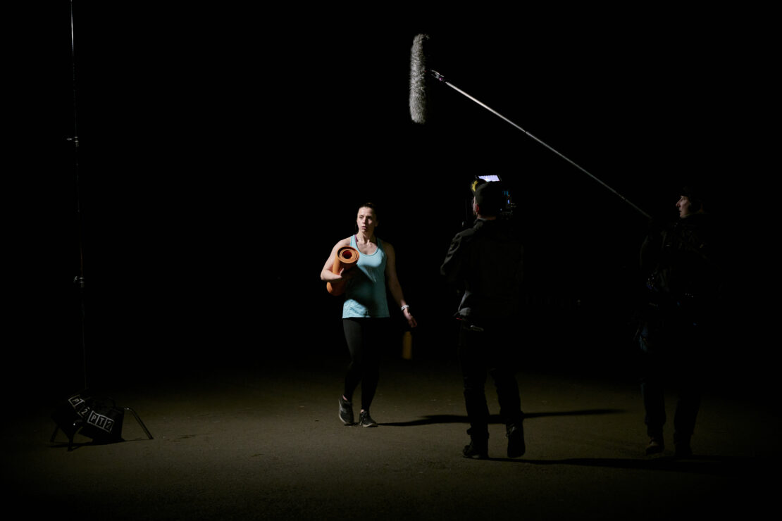 Behind the scenes of 'I'm Tired of This' film - a woman with a yoga mat walking to her car in the dark. Film and sound crew are visible.